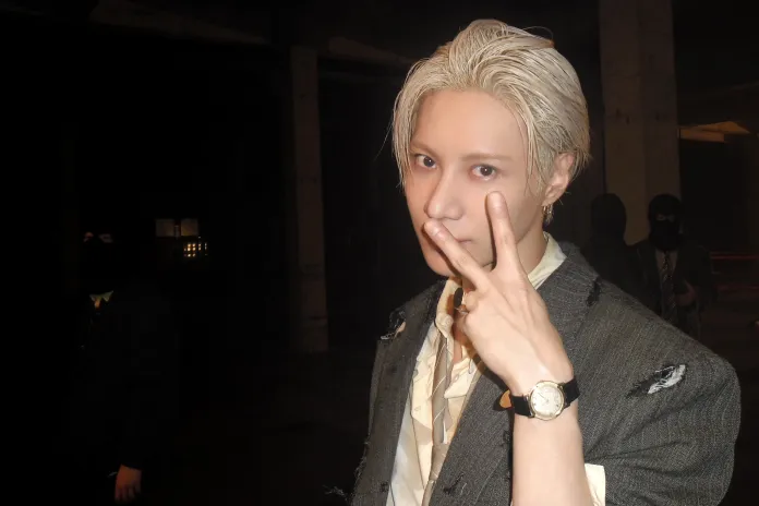 TAEMIN with light-colored hair wearing a distressed suit and a watch, holding up a peace sign in a dimly lit setting with shadowy figures in the background.