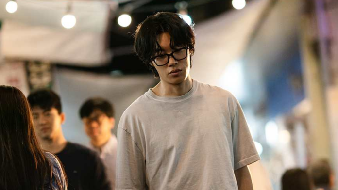 Ryu Jun Yeol in a gray t-shirt and glasses, looking down contemplatively while walking through a bustling street market with blurred figures in the background.