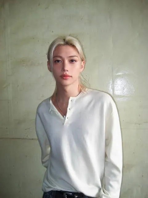 A person with long blonde hair wearing a white long-sleeve shirt, standing against a plain wall.