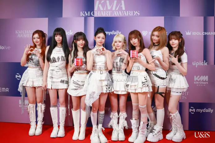 UNIS in stylish white outfits pose together at the KM Chart Awards, holding a red award and making various hand gestures, against a themed backdrop.