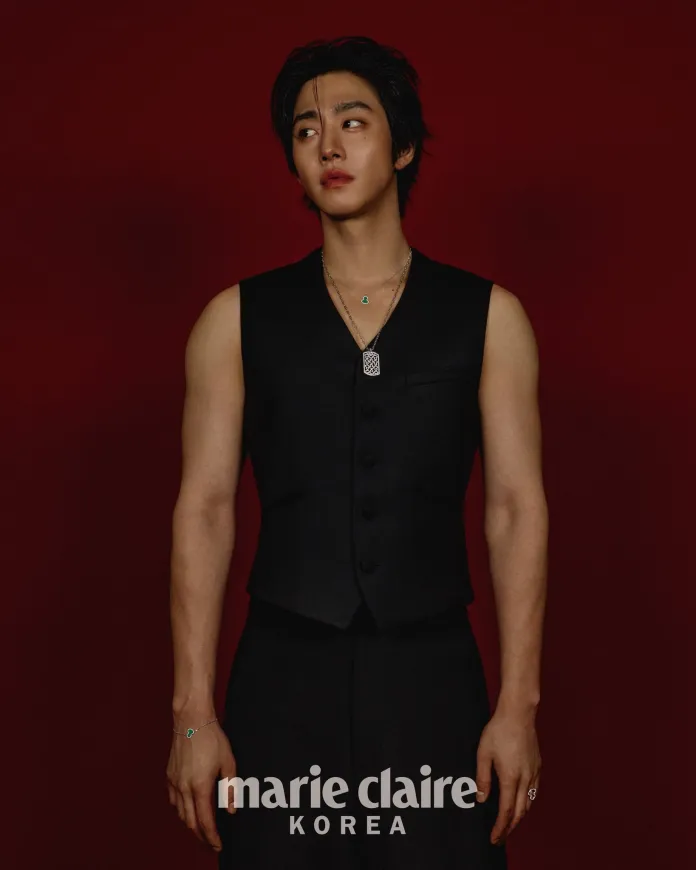 A young man in a black vest stands against a red background, looking thoughtful. He has short, styled hair and is wearing layered necklaces and jewelry.