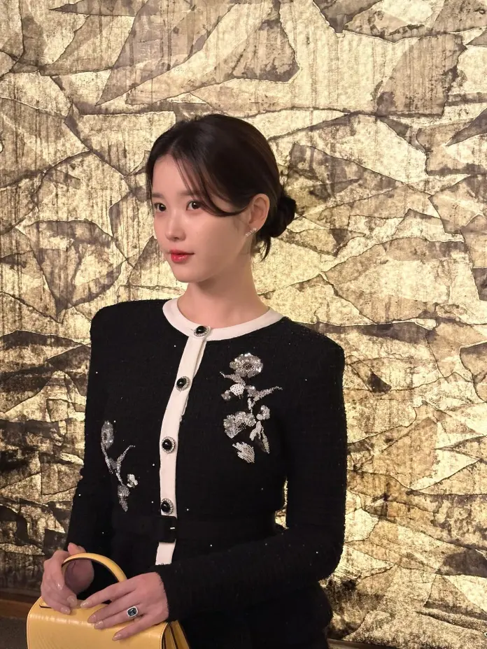 A woman in a stylish black and white outfit with floral embellishments poses against a textured golden background, holding a yellow handbag.