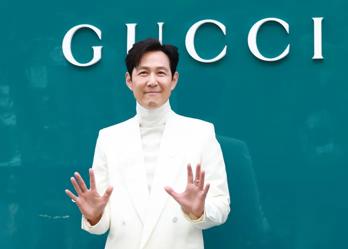Lee Jung-jae in a stylish white suit with a high collar stands in front of a green backdrop featuring the Gucci logo, gesturing with his hands in a welcoming pose.