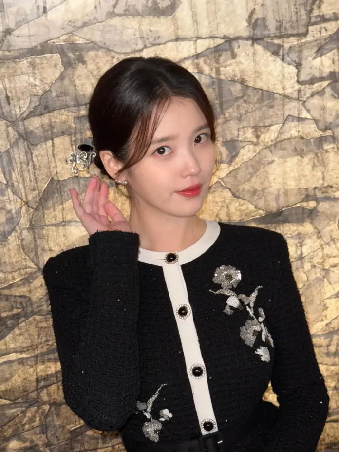 A young woman in a black and white embellished cardigan poses against a textured golden background, holding her hand to her ear.
