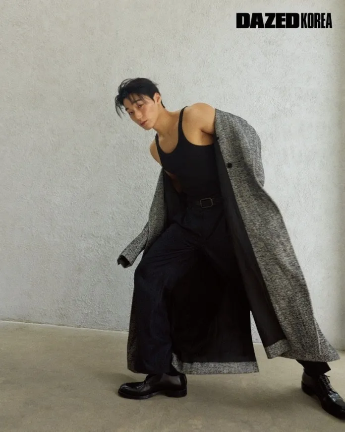 San posing in a long coat over a black tank top and dark trousers, showcasing a stylish look against a simple background.