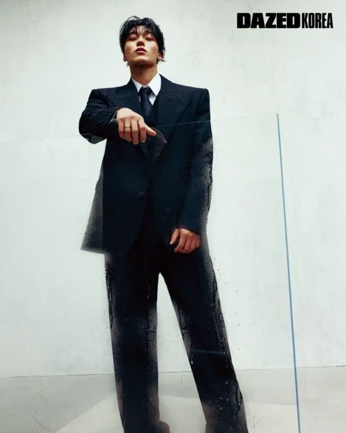 San wearing a stylish oversized black suit and tie stands confidently in a modern indoor space, with a smoky glass panel in front of them.