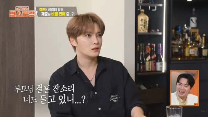 Kim Jaejoong with light brown hair sits at a table, looking surprised or concerned while engaged in conversation. His outfit is casual, featuring a dark shirt with a logo. In the background, a bar display filled with various bottles is visible. Another person is seen in a small inset, smiling.