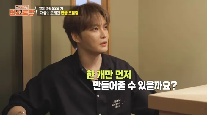 Kim Jaejoong with light brown hair, wearing a black shirt, sitting at a table and looking towards the side, appearing to be engaged in conversation.