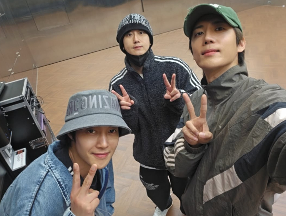Three young men posing for a selfie indoors, each making peace signs with their fingers. They are wearing casual sportswear and hats.