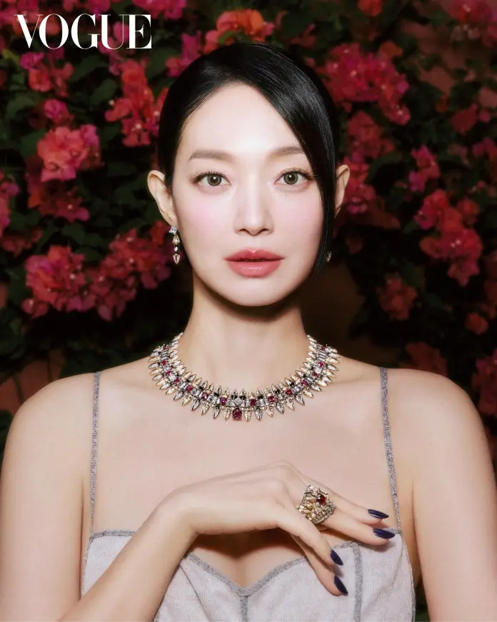 Portrait of Shin Minah wearing elegant jewelry, including a statement necklace and ring, against a vibrant floral background.
