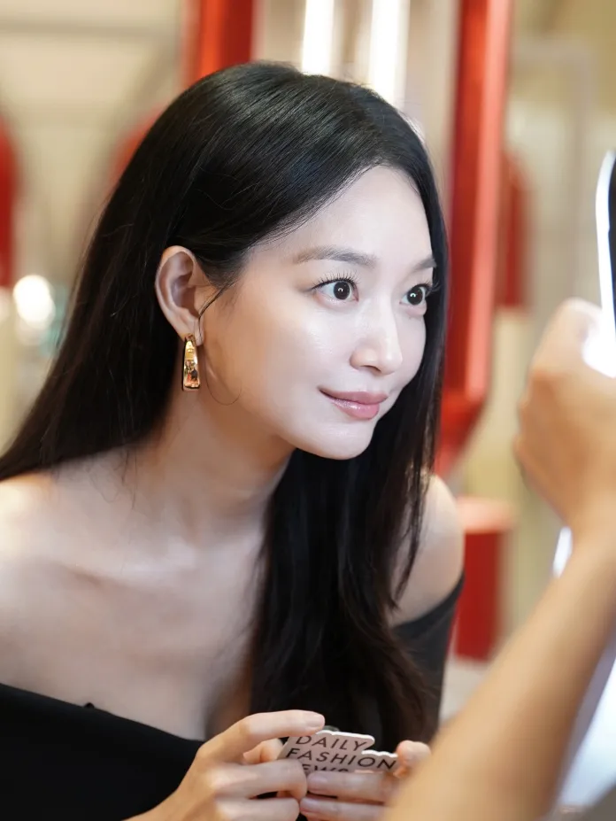 Shin Minah with long, dark hair and wearing a black off-shoulder top looks into a mirror, holding a card that says 'DAILY FASHION'.