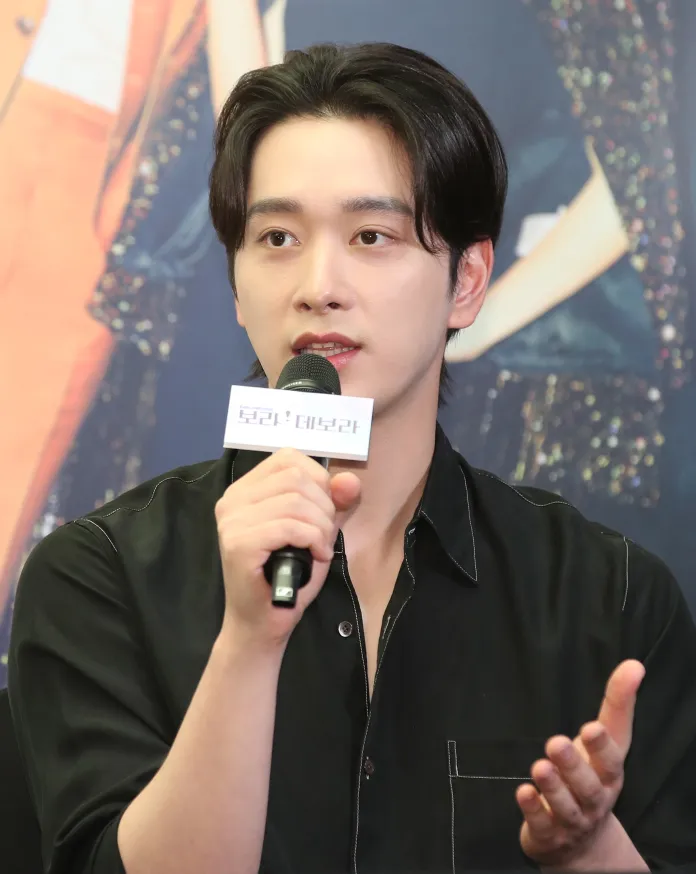 Chansung with dark hair is speaking into a microphone while holding a sign during a press conference. He is wearing a black shirt and appears engaged in conversation.