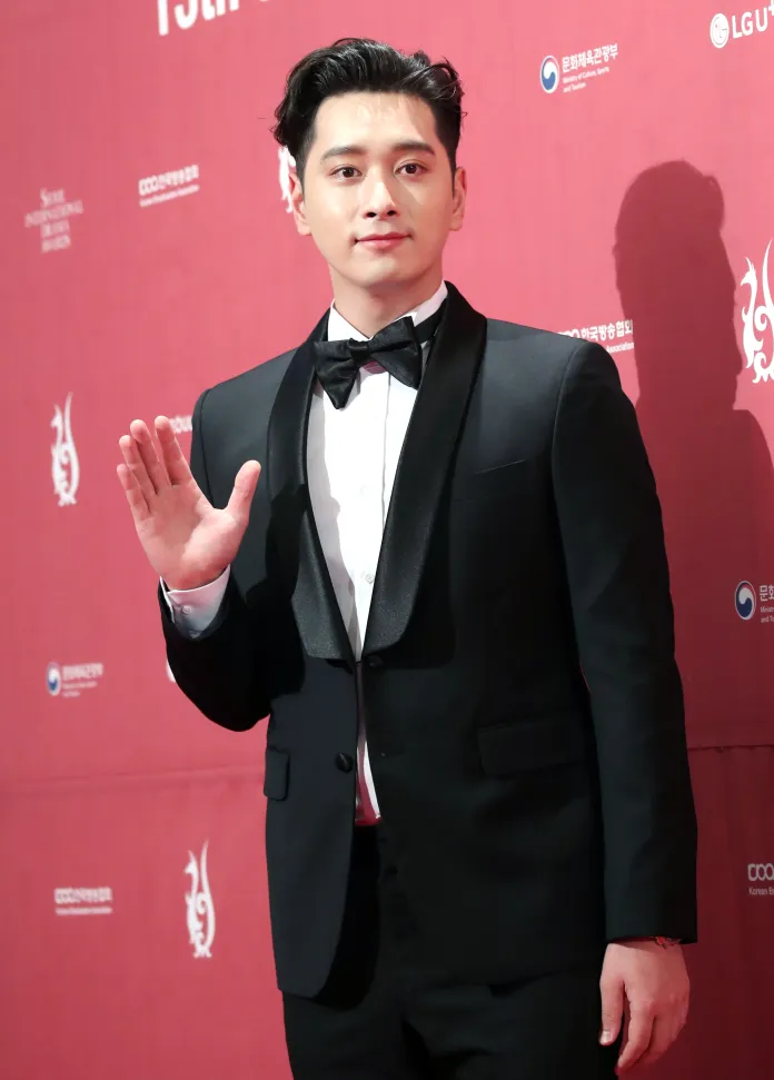 Chansung in a tuxedo waving at the camera on a red carpet event.
