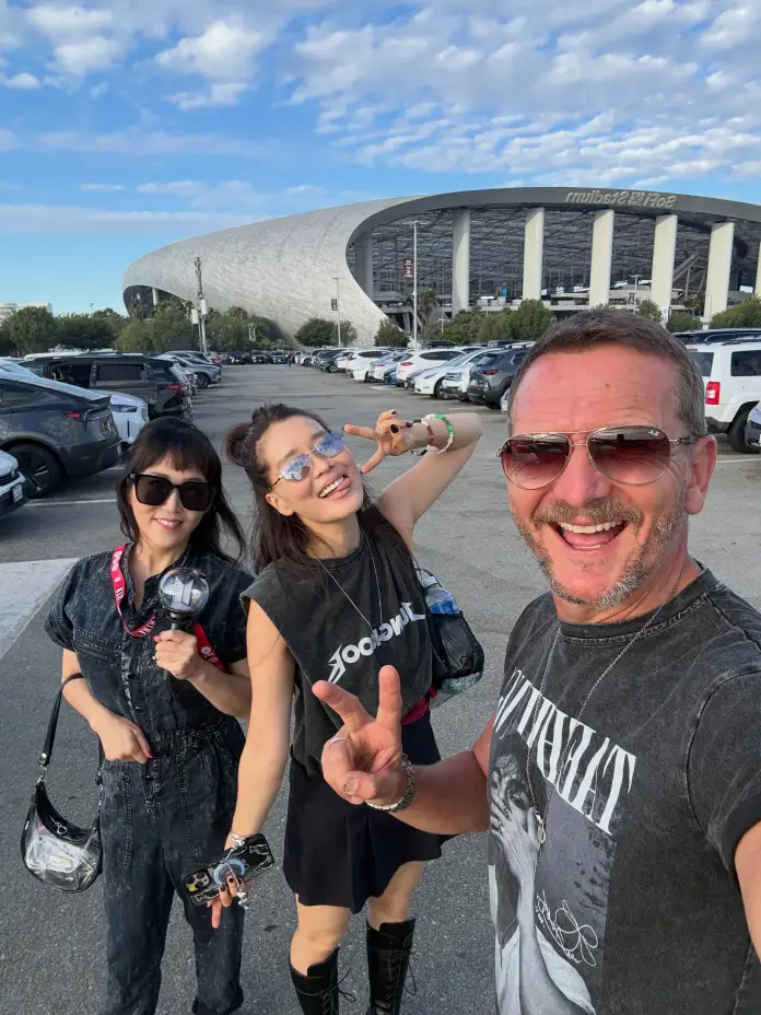 Sebastian Roché posing for a selfie in a parking lot, smiling and making peace signs, with a large modern stadium in the background.