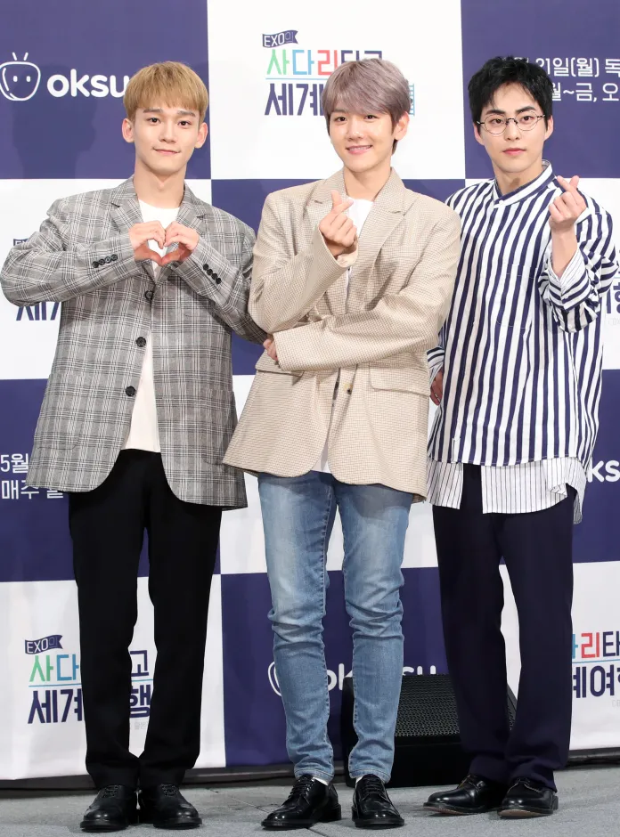 Chen, Baekhyun and Xiumin posing for a photo at an event, making hand gestures and smiling. Chen on the left is making a heart gesture, Baekhyun is showing a hand sign, and Xiumin has a playful gesture. They are dressed in stylish outfits with a patterned backdrop displaying event information.