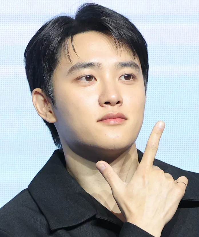 Doh Kyung Soo with short black hair and a neutral expression, making a gesture with his hand held up near his face.