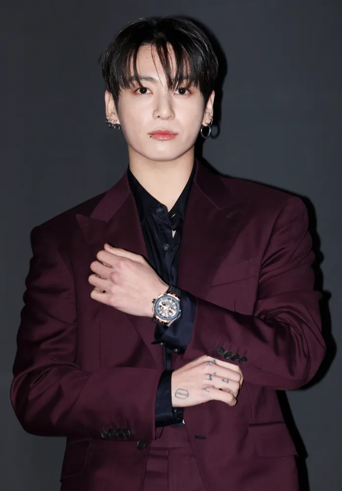 Jungkook in a maroon suit stands confidently with one hand on his arm, wearing a black shirt and a watch, against a dark background.