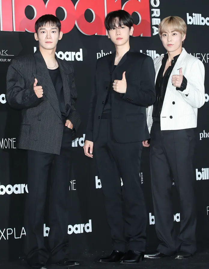 Chen, Baekhyun and Xiumin posing for a photo at a Billboard event, each giving a thumbs up. They are dressed in stylish outfits, with Chen wearing a striped jacket, Baekhyun in a black suit, and Xiumin in a white blazer.