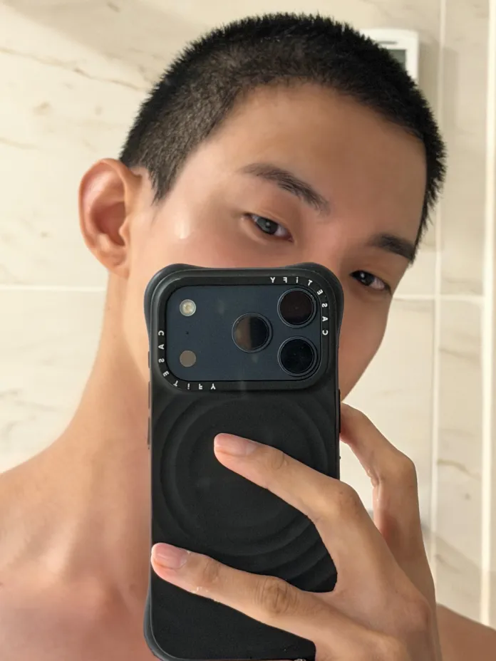 A close-up selfie of a person with short hair, holding a smartphone in front of their face in a bathroom setting.