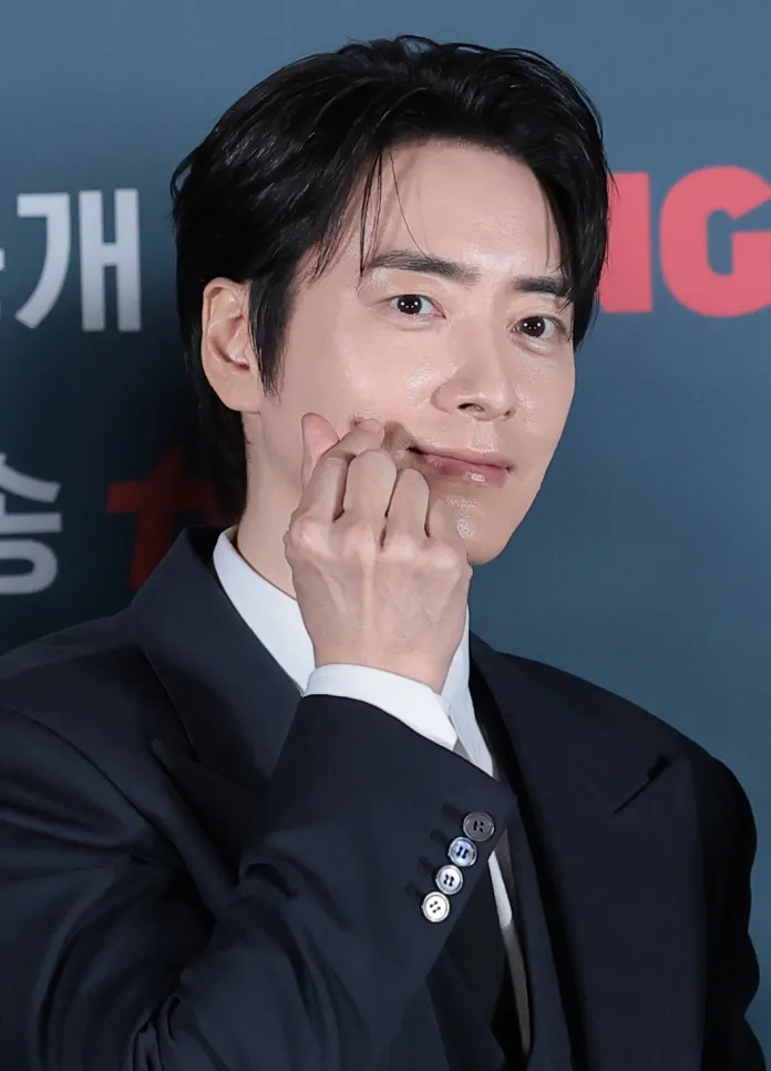 Lee Joon Hyuk in a dark suit poses with a subtle smile, touching their face with one hand against a blue backdrop.