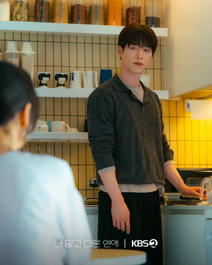 Seo Kang-joon in a gray sweater stands in a modern kitchen, looking towards a seated woman. The kitchen features shelves with jars and cups, and a warm yellow light illuminates the scene.