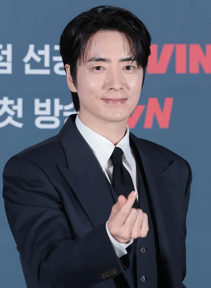 Lee Joon Hyuk smiling at the camera while making a hand gesture commonly known as a finger heart.