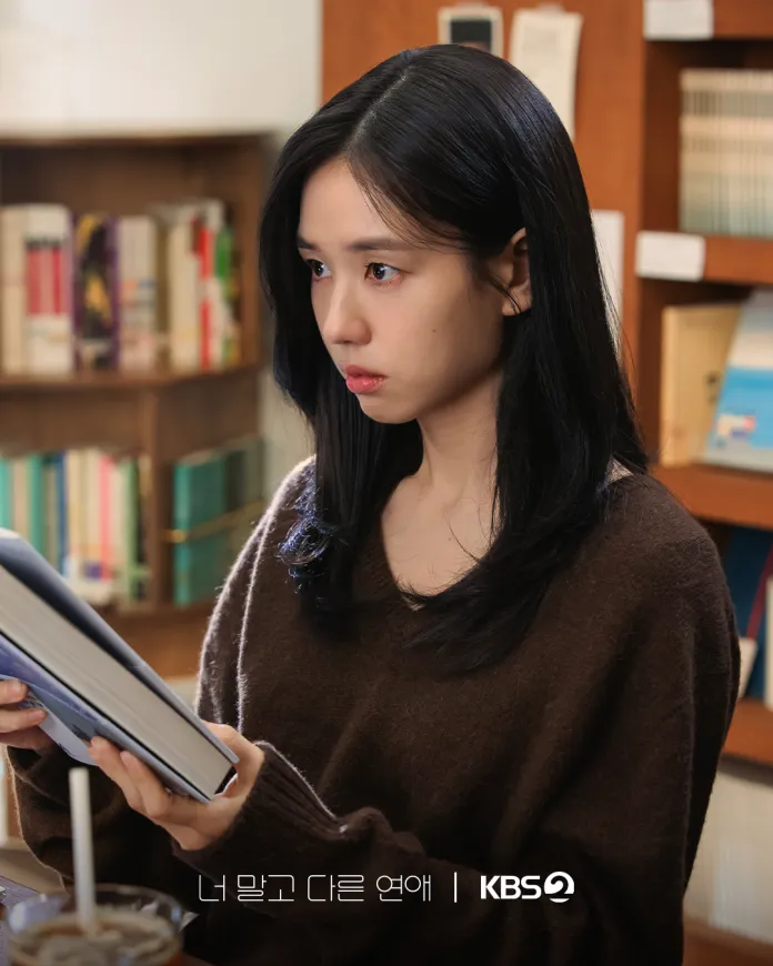 Ahn Eun-jin with long black hair is sitting in a cozy library, holding an open book and looking thoughtfully to the side. She is wearing a brown sweater, and bookshelves filled with books are visible in the background.