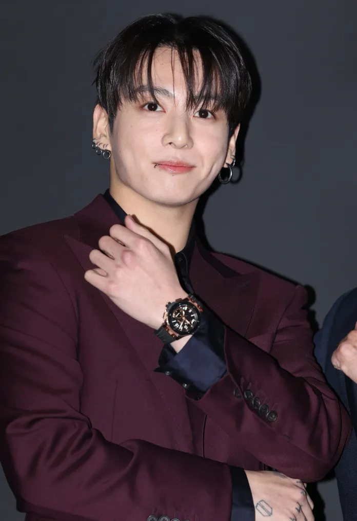 Jungkook in a burgundy suit poses with a smile, showcasing a stylish watch on his wrist, against a dark background.