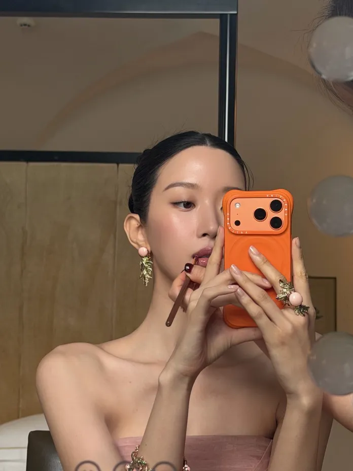 Moon Ga-young in a strapless pink dress using a makeup brush while holding a bright orange phone in front of a mirror.