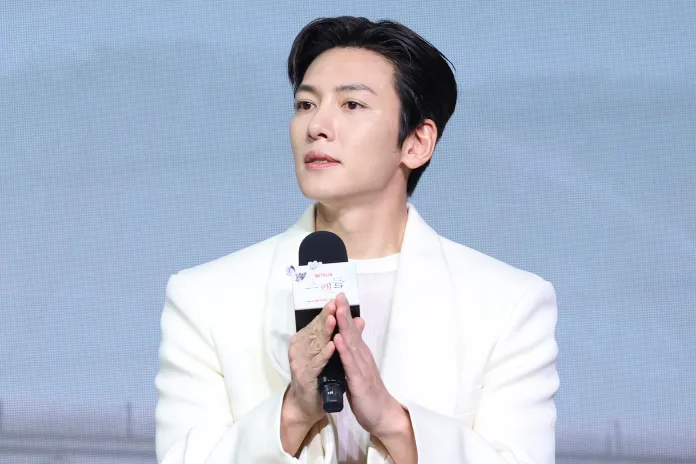 A man in a white suit is speaking into a microphone at a public event, expressing a thoughtful demeanor.