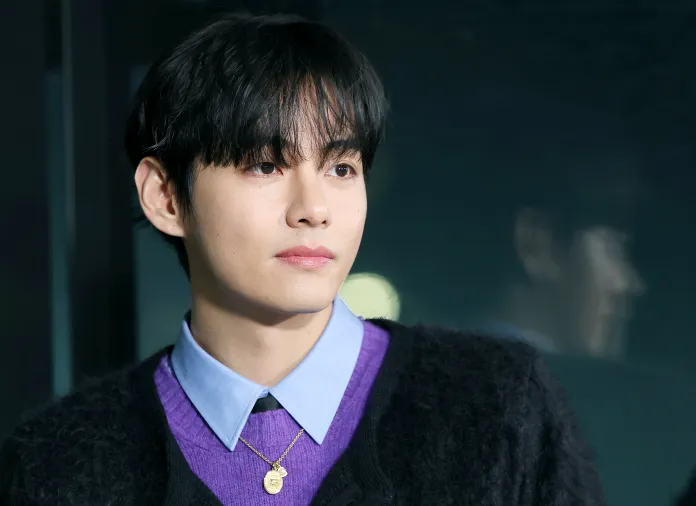 V with dark, tousled hair, wearing a purple sweater and light blue collared shirt, looking thoughtfully off-camera with a blurred reflection in the background.