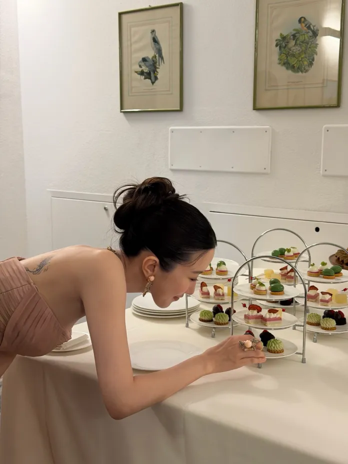 Moon Ga-young in a blush-colored dress is reaching for a dessert from a multi-tiered display of pastries and sweets, with decorative plates in the background.