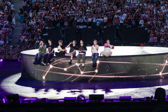 BTS sitting on a circular stage during a concert, facing an audience. The stage is illuminated with colorful lights, and a large crowd can be seen in the background, cheering.