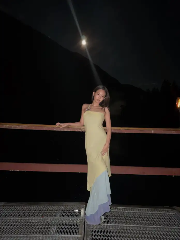 Jennie stands on a metal platform at night, wearing a long dress that is yellow at the top and transitions to soft pastel colors at the bottom. The moon is visible in the background, illuminating the scene.
