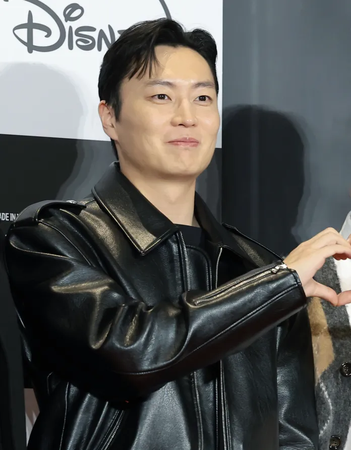 Noh Jae-won wearing a black leather jacket making a heart shape with his hands, smiling at an event with a Disney backdrop.