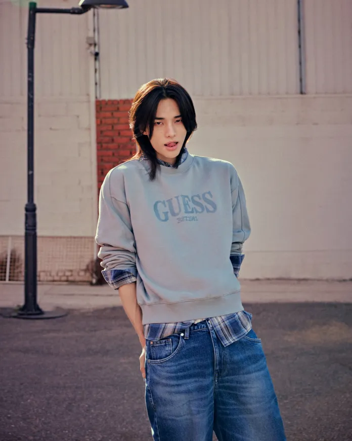 Hyunjin standing outdoors in front of a wall, wearing a light blue Guess sweatshirt layered over a plaid shirt, paired with blue jeans. The lighting casts a warm tone on the scene.