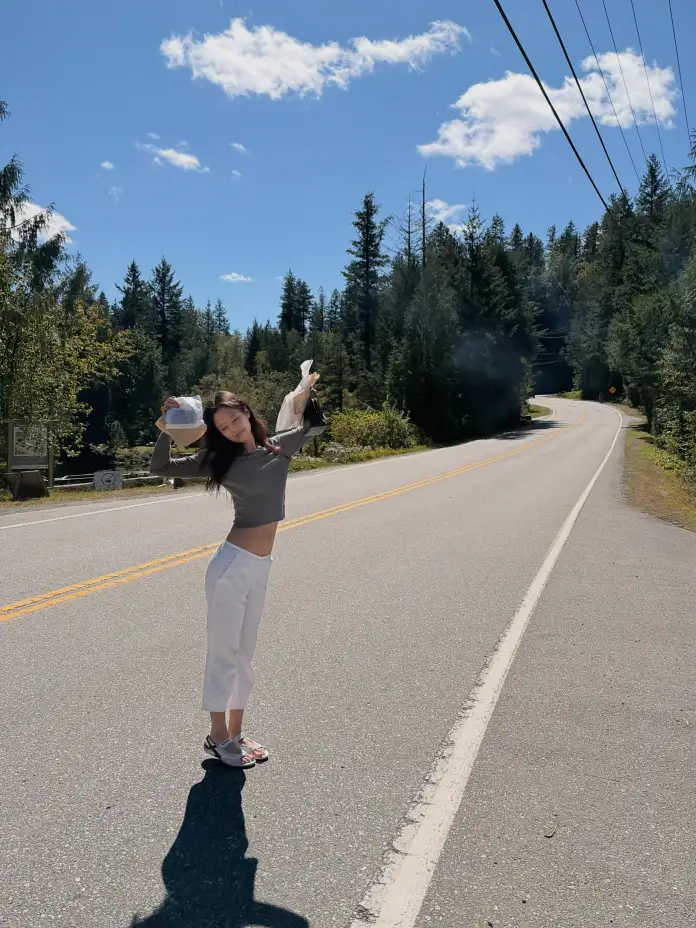 Jennie standing on the roadside with her arms raised, surrounded by trees and a clear sky.