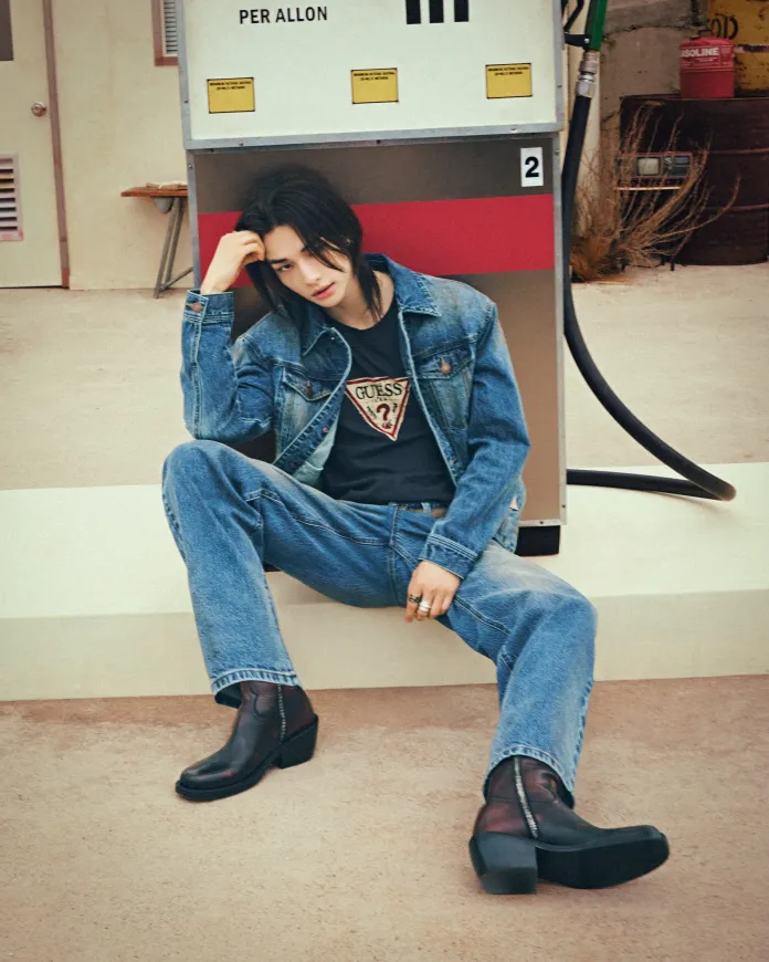 Hyunjin sitting casually in a denim jacket and jeans, wearing a black t-shirt with a logo, black and red ankle boots, posed in front of a gas station backdrop.