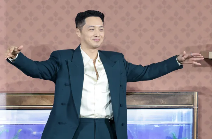 Byun Yo-han in a dark suit and light shirt smiles while extending his arms open wide, standing in front of a decorative background.
