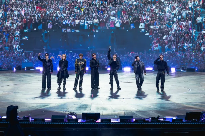 BTS on stage, dressed in stylish outfits, waving to a cheering crowd at a large concert. Bright lights illuminate the stage, with fans visible in the background.