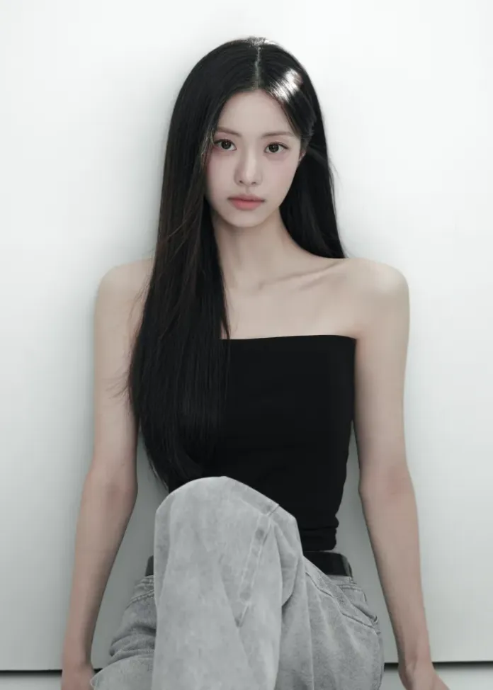 A portrait of a young woman sitting against a white wall, wearing a strapless black top and light gray jeans, with long black hair cascading down her shoulders.