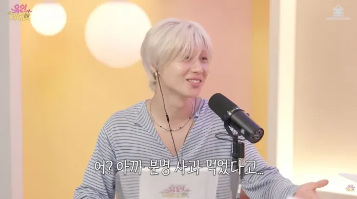Taemin with light-colored hair wearing a striped shirt smiles while speaking into a microphone in a brightly lit room.
