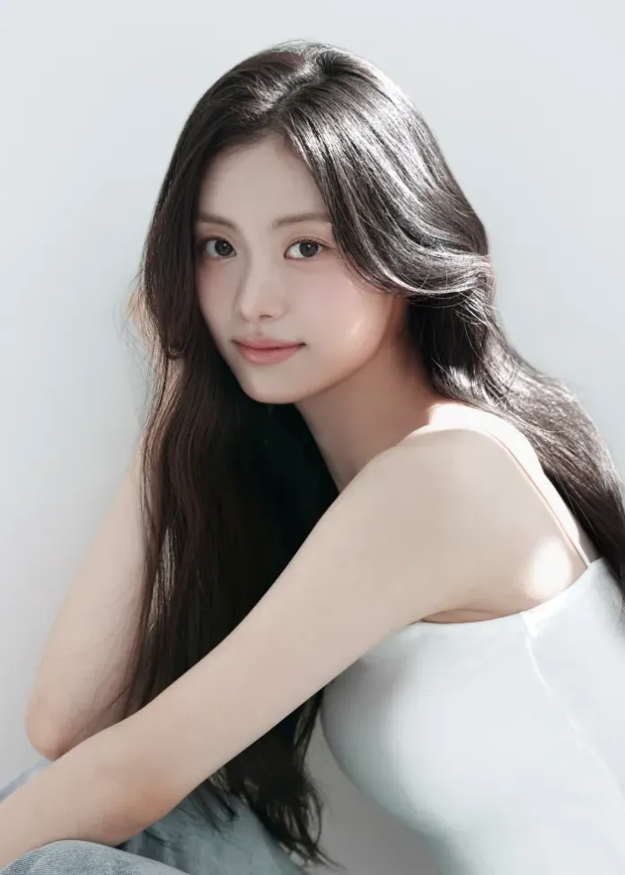 A young woman with long, dark hair poses softly with a smile, wearing a sleeveless white top against a light background.