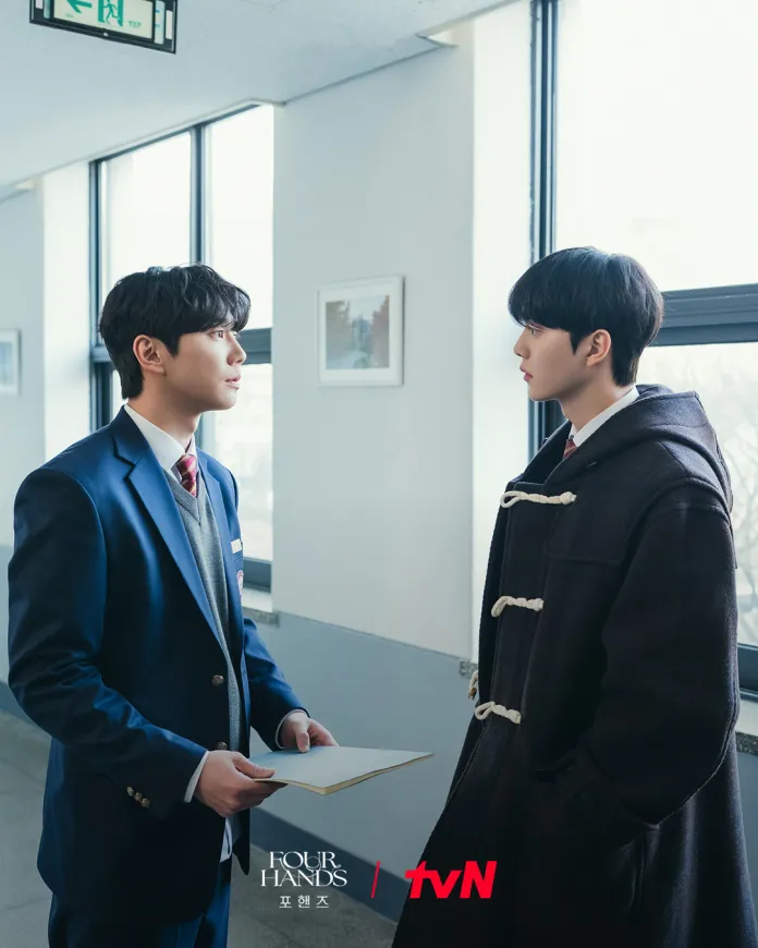Song Kang and Lee Junyoung in a hallway, one in a blue suit holding a folder, while the other, in a black coat, stands facing him. Sunlight comes through large windows.
