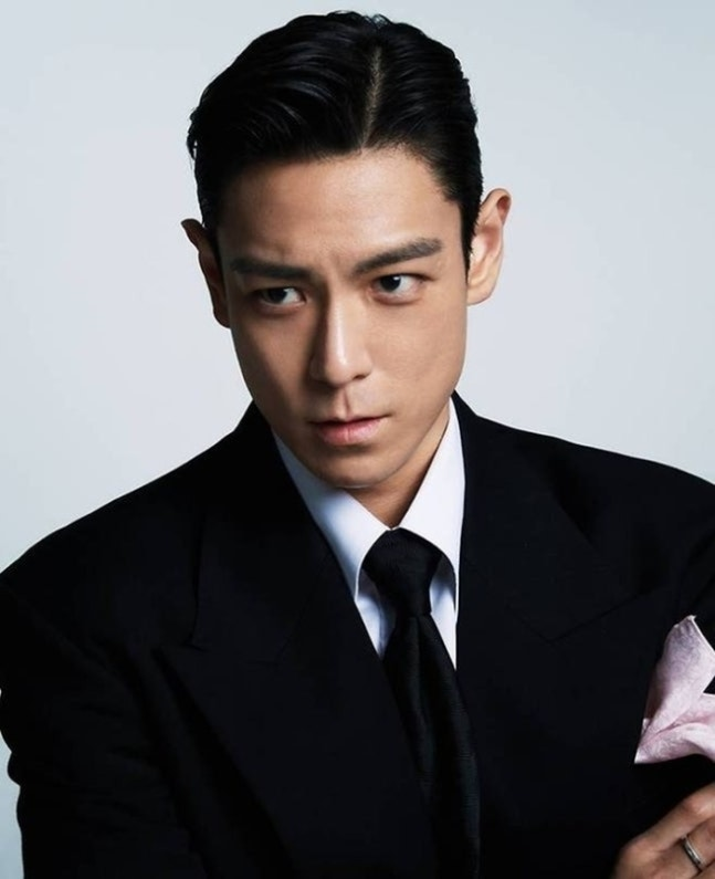 A young man in a classic black suit with a white shirt and black tie, giving a serious expression. He has slicked-back hair and is accessorized with a pink handkerchief tucked into his suit pocket.
