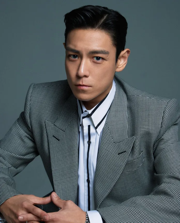 Close-up portrait of a man in a stylish grey checkered suit, looking seriously at the camera with his hands resting on a table.