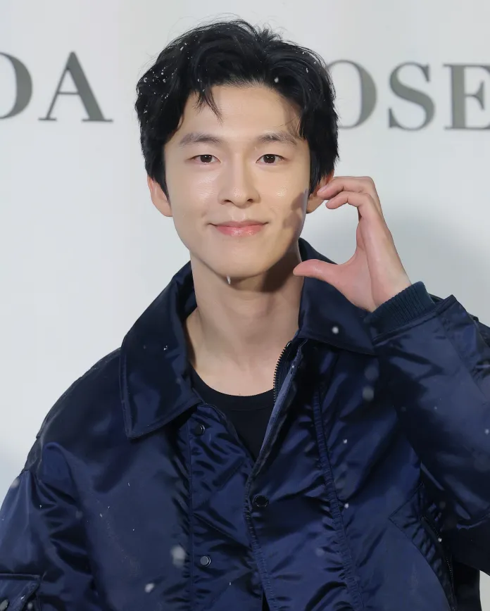 Hong Kyung smiling and posing with his hand near his face, wearing a dark blue jacket, with snowflakes falling around him.
