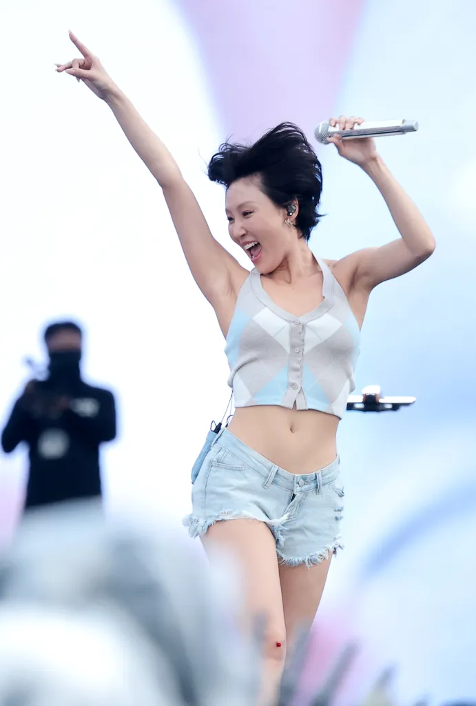 Hwasa on stage energetically singing with a microphone in hand, wearing a sleeveless top and denim shorts, while raising one arm in excitement.
