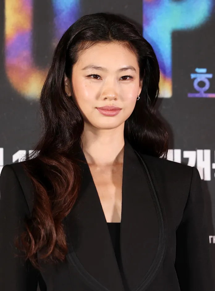 Hoyeon with long, wavy black hair wearing a black suit, smiling at the camera against a colorful backdrop.