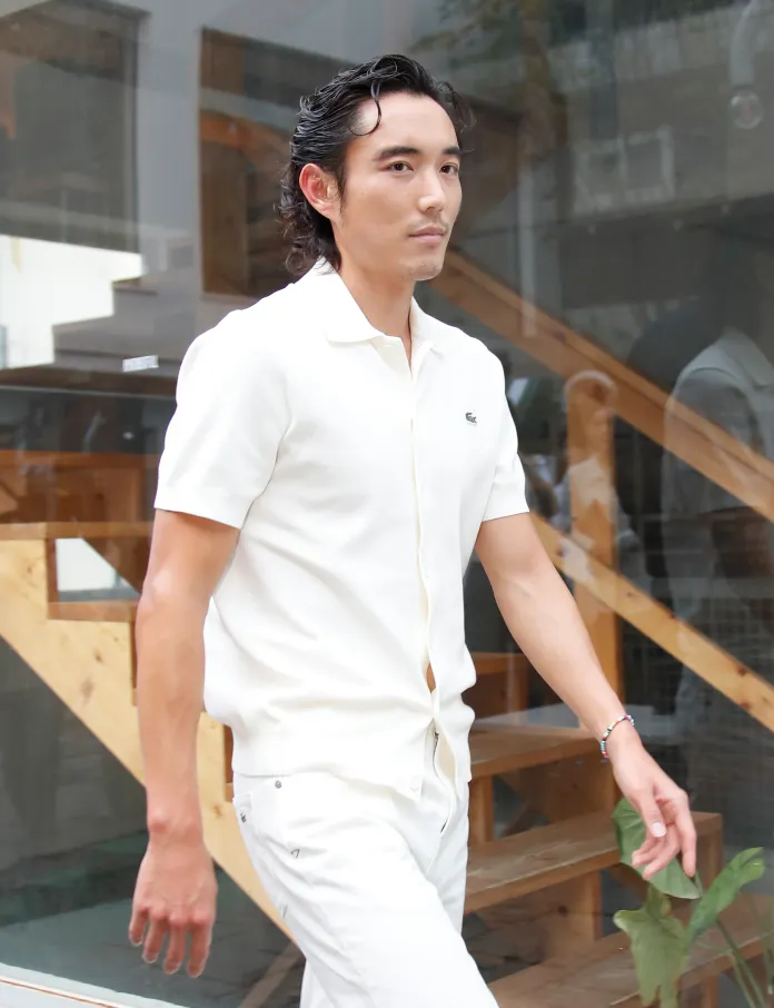 Justin H. Min with dark, wavy hair wearing a white short-sleeve shirt and white pants, walking past a wooden staircase and glass wall.